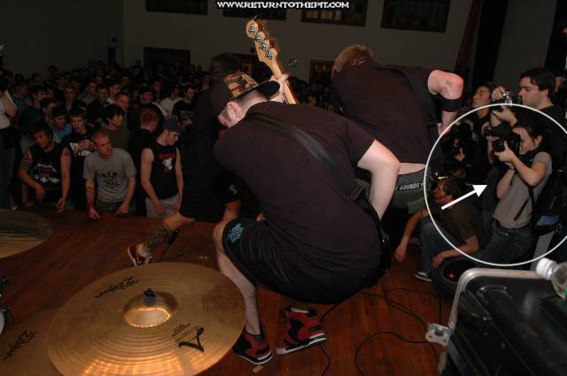 Two guitarists jump in the air while a small person off to the side photographs.