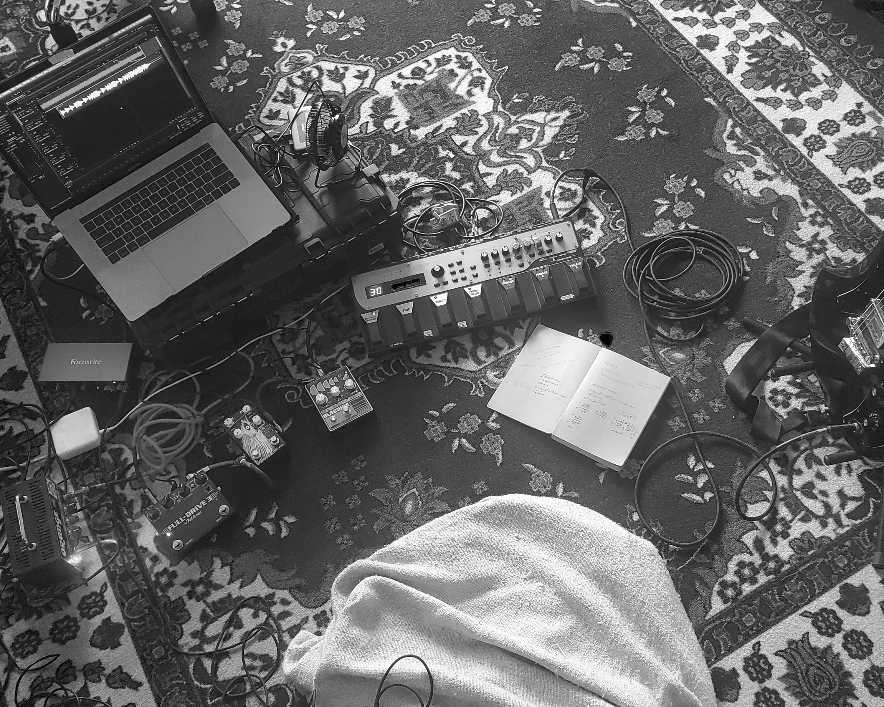 Guitar pedals, a laptop, and an open journal spread out over a carpet