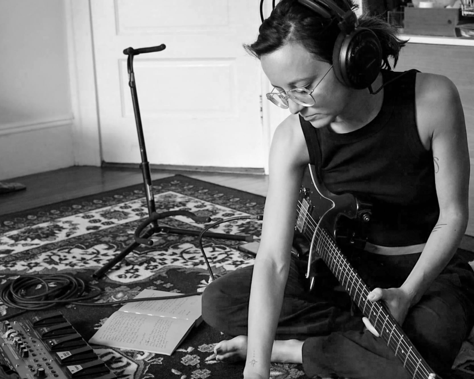 The artist sitting on the floor holding a guitar in one hand and reaching towards something in the foreground with the other.