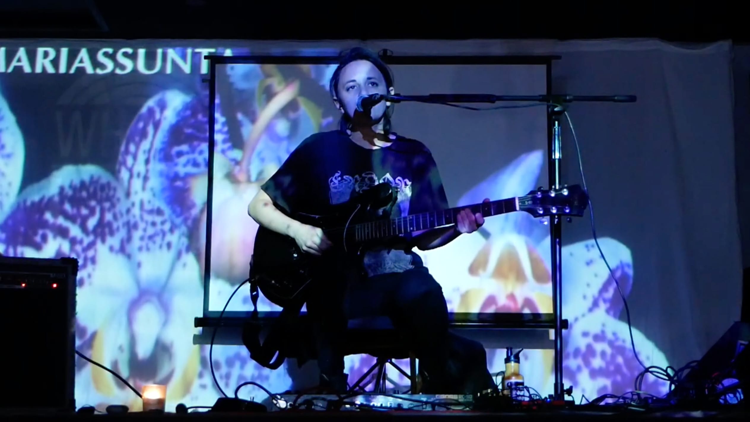 Mariassunta playing an electric guitar live, with a projected image of a purple flower flooding the stage